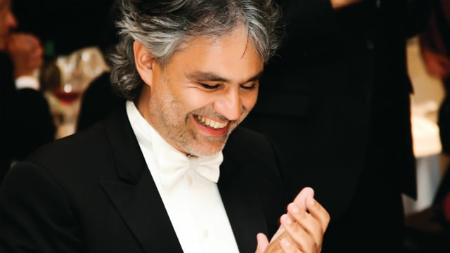 Primo piano di Andrea Bocelli in smoking scuro e papillon bianco. Il cantante applaude sorridente.