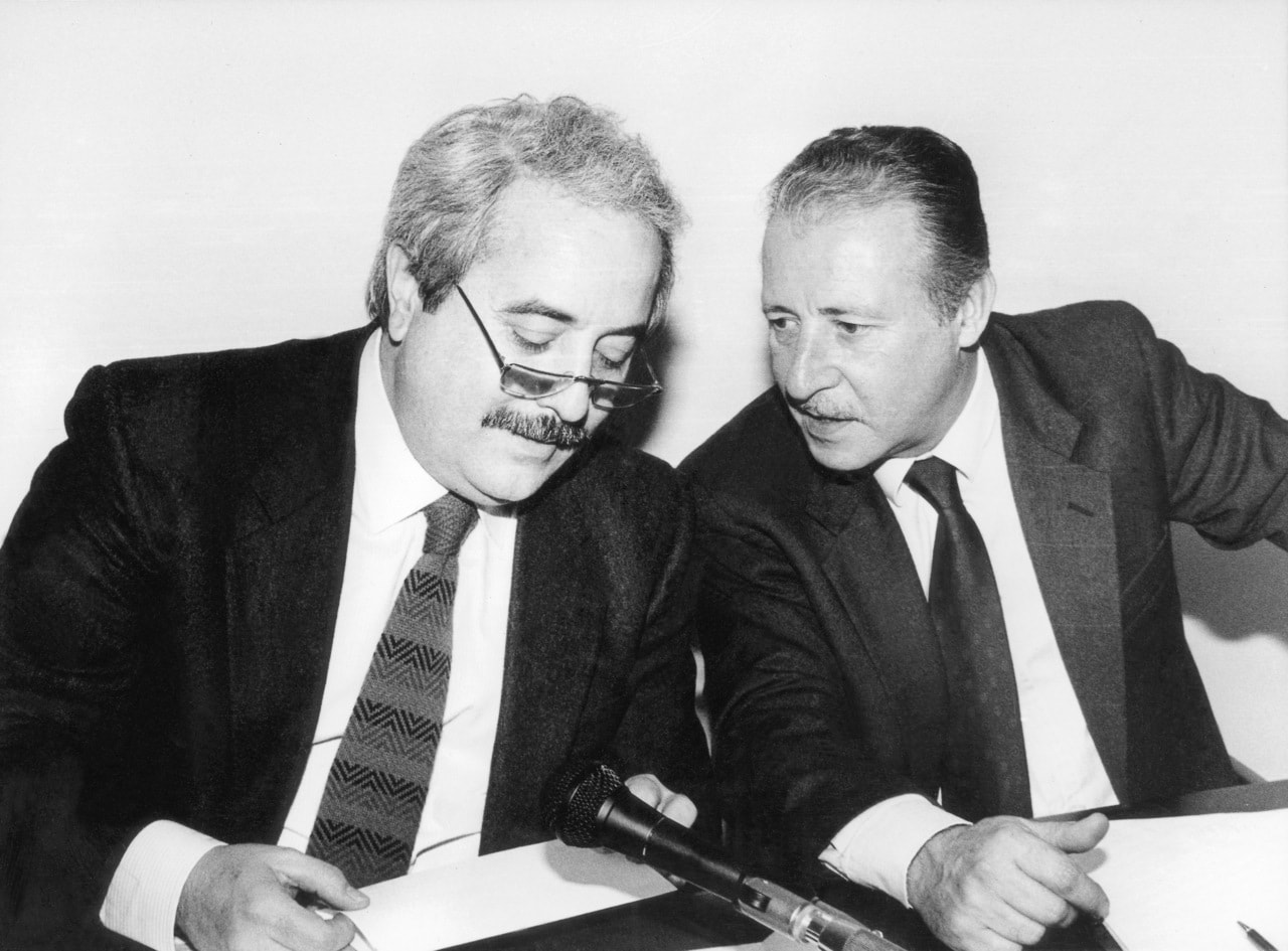 Foto in bianco e nero che ritrae Paolo Borsellino che sussurra qualcosa a Giovanni Falcone mentre siedono uno accanto all'altro.