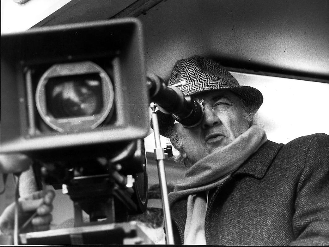 Foto in bianco e nero di Fellini dietro la macchina da presa. Il regista indossa cappotto, la tipica sciarpa e cappello in tessuto principe di Galles dal quale fuoriescono ciuffi di capelli brizzolati.
