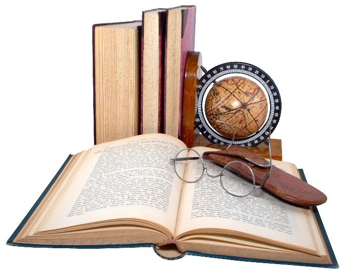 Un dizionario aperto su sfondo bianco, sopra due occhiali e un porta occhiali, sullo sfondo un piccolo mappamondo e altri libri.