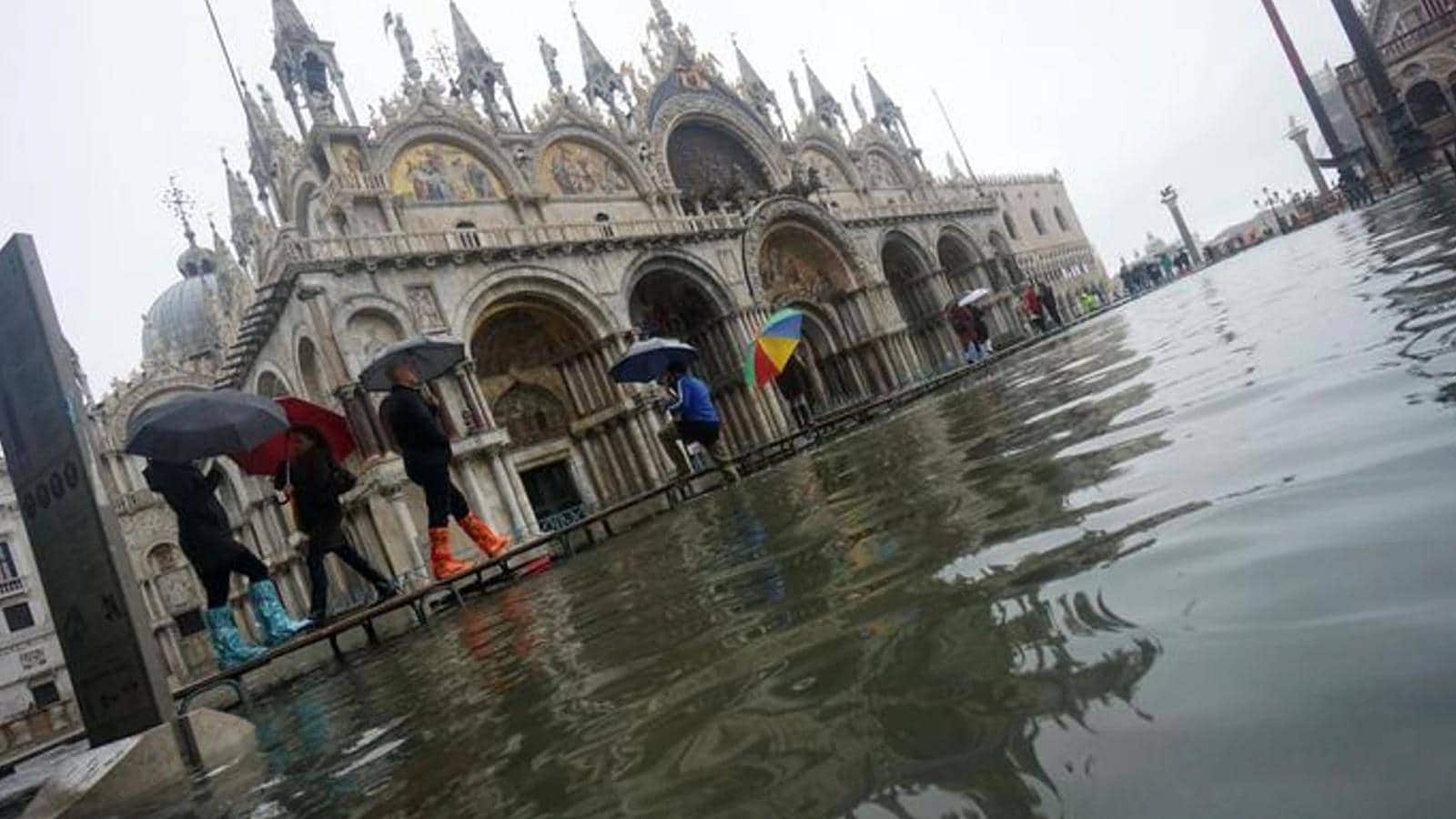 Acqua alta in piazza San Marco: cittadini e turisti camminano sulla passerella con calosce ai piedi e ombrelli aperti.