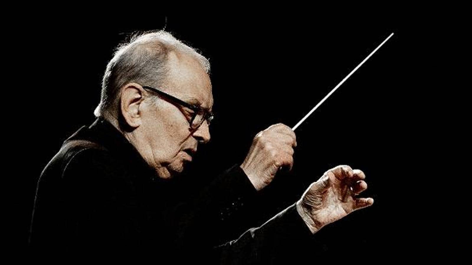 Primo piano di profilo del Maestro Ennio Morricone, con giacca nera, fotografato su sfondo nero mentre dirige. La luce illumina il suo volto concentrato e le sue mani, bacchetta nella destra, mentre guidano un'orchestra.