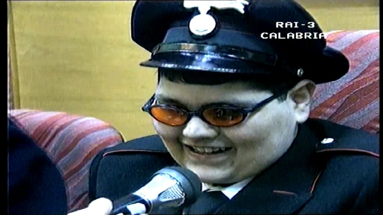Primo piano di Piero sorridente, con la divisa da Carabiniere e gli occhiali da sole, mentre viene intervistato dalla giornalista della TGR Calabra.