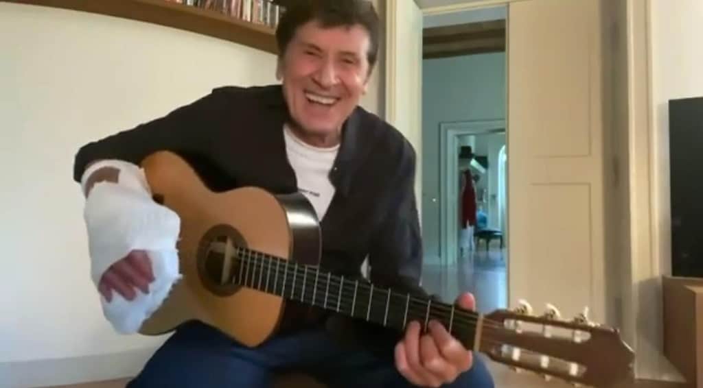 Gianni Morandi suona la chitarra con la mano fasciata, ride e intona